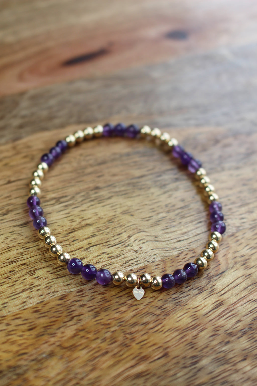 A beaded stretch bracelet featuring purple amethyst beads and polished gold spacer beads, with a small gold heart charm, resting on a wooden surface.