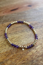 A beaded stretch bracelet featuring purple amethyst beads and polished gold spacer beads, with a small gold heart charm, resting on a wooden surface.
