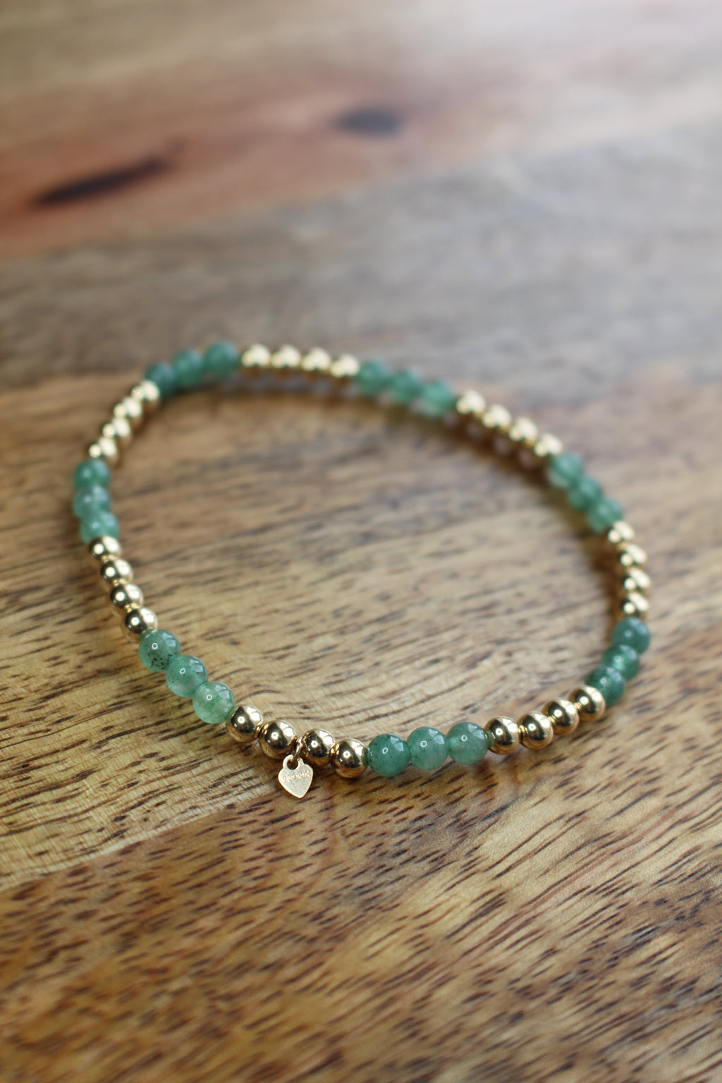 A beaded stretch bracelet featuring green aventurine beads and small gold-toned spacer beads, with a tiny gold heart charm, resting on a wooden surface.