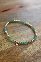 A beaded stretch bracelet featuring green aventurine beads and small gold-toned spacer beads, with a tiny gold heart charm, resting on a wooden surface.