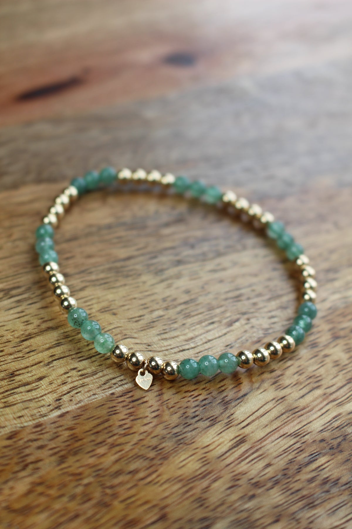 A beaded stretch bracelet featuring green aventurine beads and small gold-toned spacer beads, with a tiny gold heart charm, resting on a wooden surface.