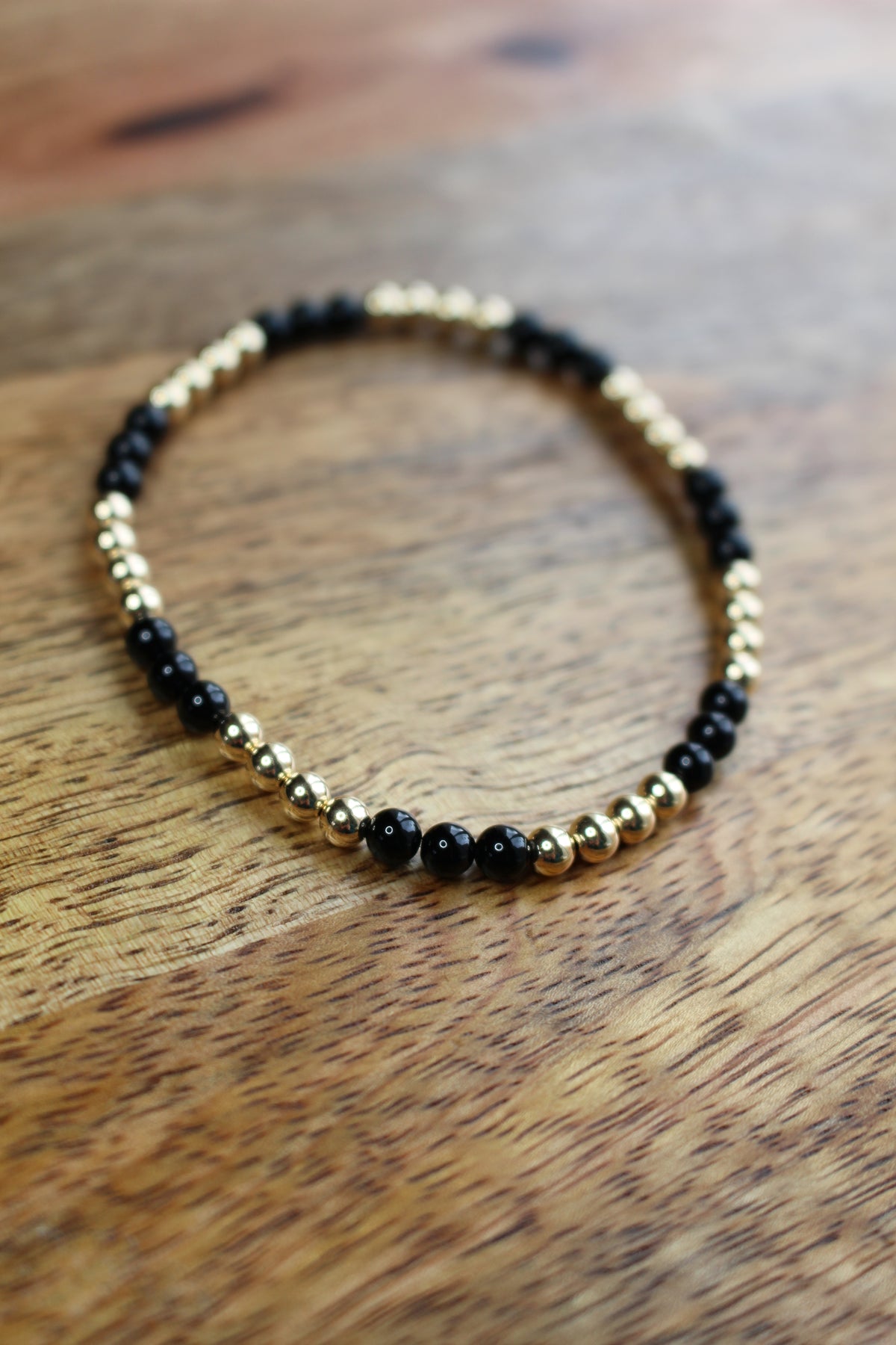 A beaded stretch bracelet with alternating black onyx and gold metallic beads, displayed on a warm brown wooden surface.