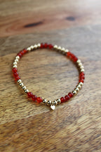 A beaded stretch bracelet featuring alternating translucent red Carnelian beads and polished 14ct gold filled metallic beads, with a small gold heart charm at the front, displayed on a wooden surface.