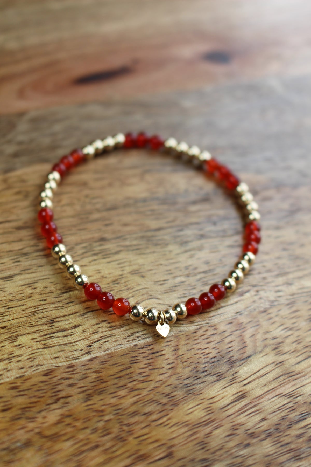A beaded stretch bracelet featuring alternating translucent red Carnelian beads and polished 14ct gold filled metallic beads, with a small gold heart charm at the front, displayed on a wooden surface.