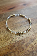 A beaded stretch bracelet with gold-toned metal beads and clear quartz crystal beads, resting on a rustic wooden surface.