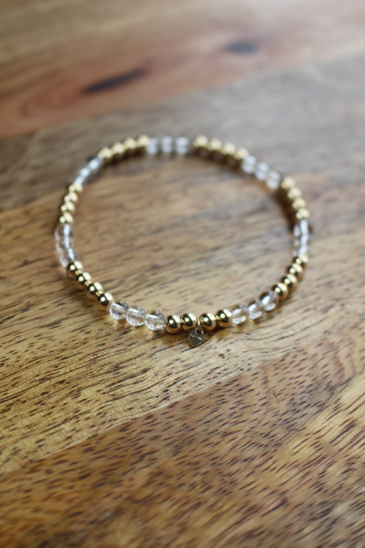 A beaded stretch bracelet with gold-toned metal beads and clear quartz crystal beads, resting on a rustic wooden surface.