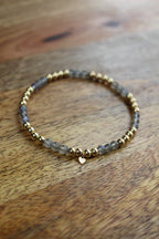 A beaded stretch bracelet featuring grey labradorite stones and gold-toned spacer beads, with a small gold heart charm, resting on a wooden surface.
