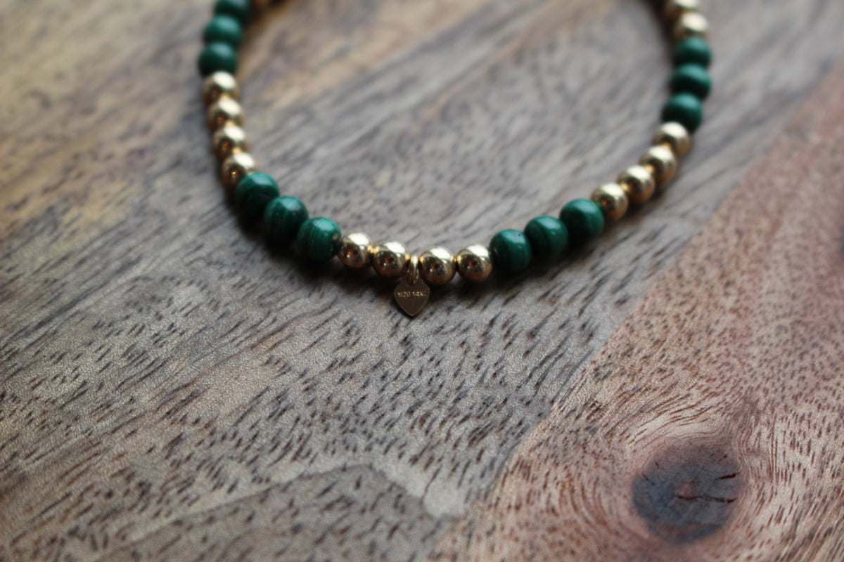 A beaded bracelet with dark green malachite and metallic gold beads, featuring a small heart-shaped charm engraved with 14ct filled', resting on a rustic wooden surface.