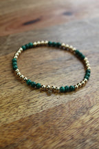 A beaded stretch bracelet featuring dark green malachite beads and polished gold spacer beads, resting on a rustic wooden surface.