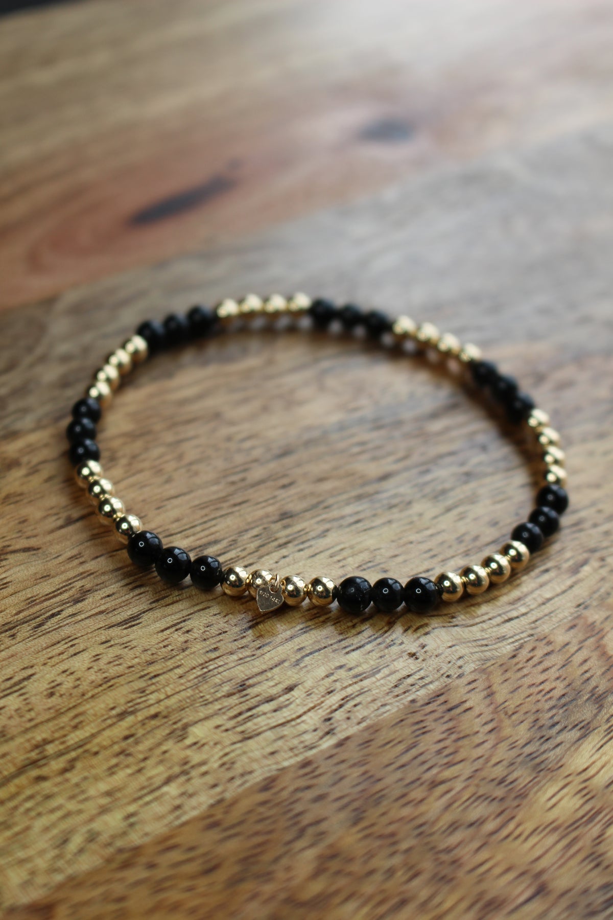 A beaded stretch bracelet with alternating Obsidian and gold metallic beads, featuring a small silver heart charm in the center, displayed on a wooden surface.