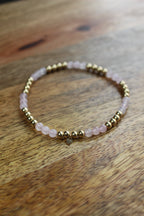 A beaded stretch bracelet featuring light pink rose quartz beads and polished gold spacer beads, resting on a rustic wooden surface.