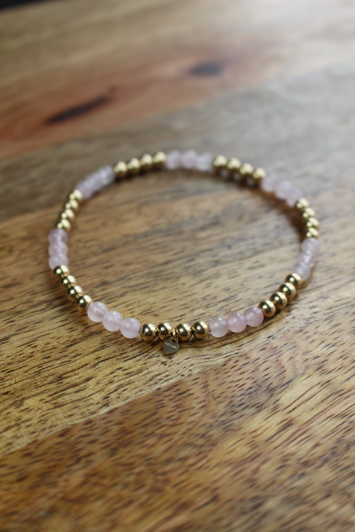 A beaded stretch bracelet featuring light pink rose quartz beads and polished gold spacer beads, resting on a rustic wooden surface.