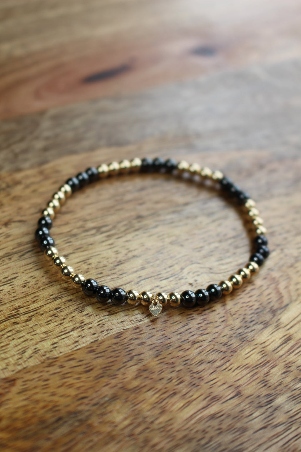 A beaded stretch bracelet with alternating black Hematite and gold metallic beads, featuring a small clear crystal charm, displayed on a wooden surface.