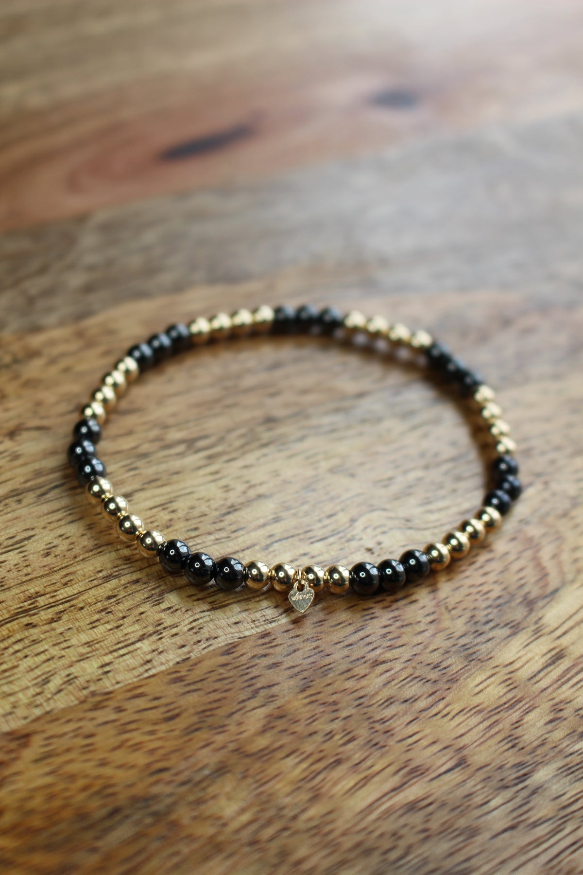 A beaded stretch bracelet with alternating black Hematite and gold metallic beads, featuring a small clear crystal charm, displayed on a wooden surface.