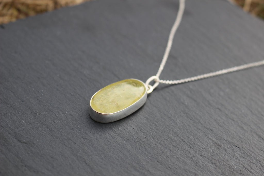 A silver oval pendant necklace with a pale yellow-green stone, resting on a dark slate surface.