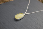 A silver oval pendant necklace with a pale yellow-green stone, resting on a dark slate surface.