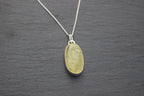 A silver chain necklace with an oval pendant featuring a textured, marbled yellow-green stone set in a silver bezel, displayed on a dark gray slate background.