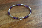 A beaded stretch bracelet featuring purple amethyst beads and polished silver-toned spacer beads, with a small silver heart charm, displayed on a wooden surface.