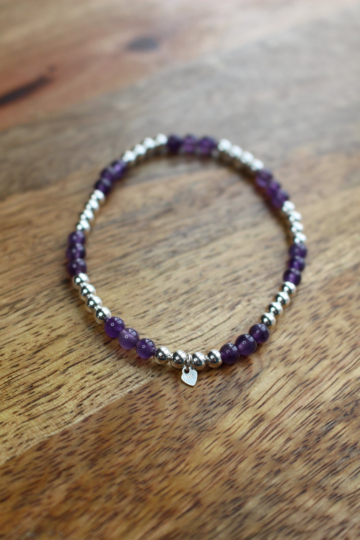 A beaded stretch bracelet featuring purple amethyst beads and polished silver spacer beads, with a small silver heart charm, displayed on a wooden surface.