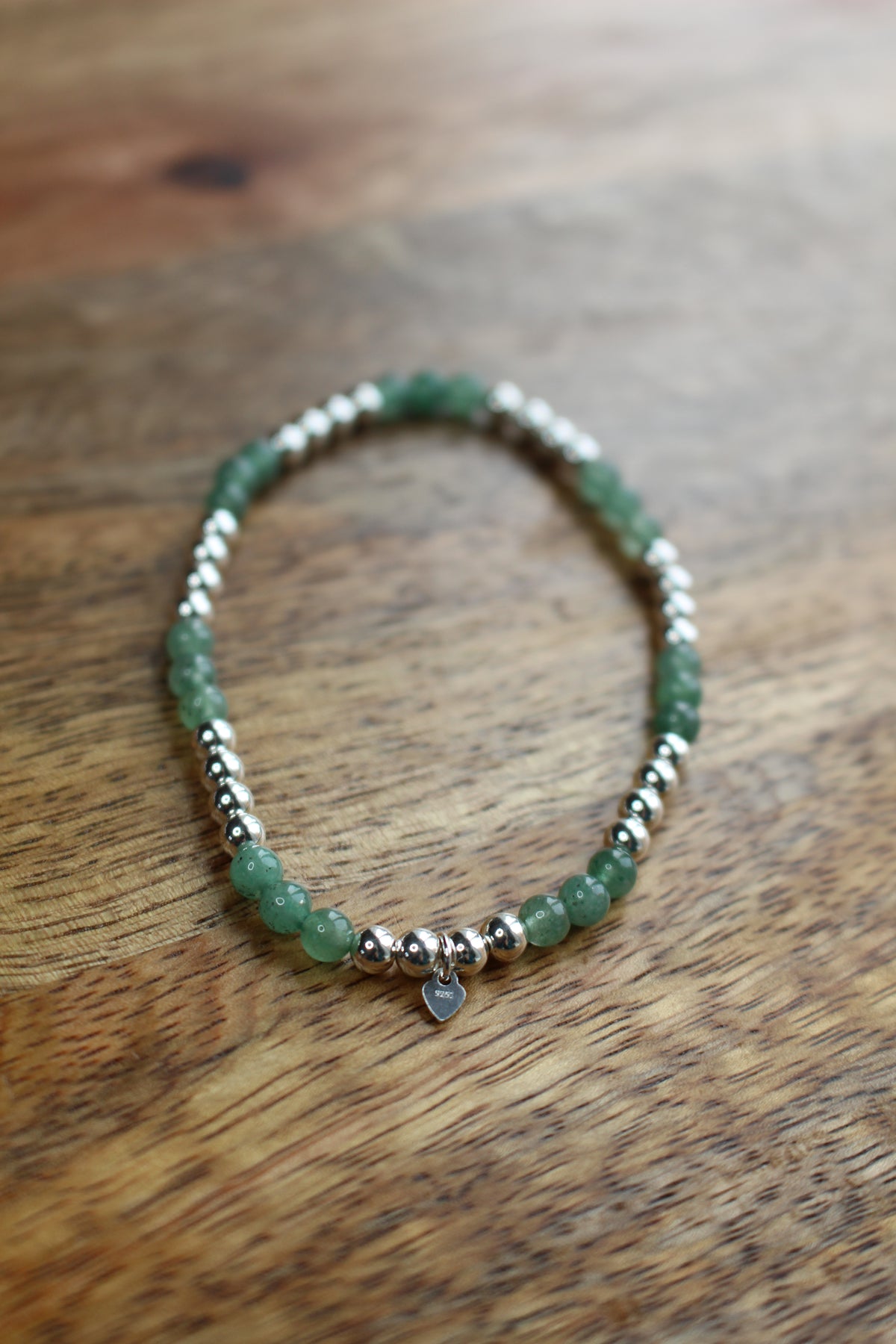 A beaded stretch bracelet with green aventurine faceted beads, silver spacer beads, and a small silver heart charm, displayed on a wooden surface.