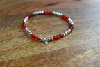 A beaded stretch bracelet with alternating translucent red Carnelian and polished silver beads, featuring a small silver heart charm, displayed on a wooden surface.