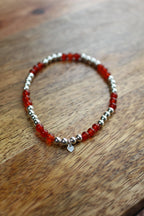 A beaded stretch bracelet featuring translucent red carnelian beads and polished silver spacer beads, with a small silver heart charm, displayed on a wooden surface.