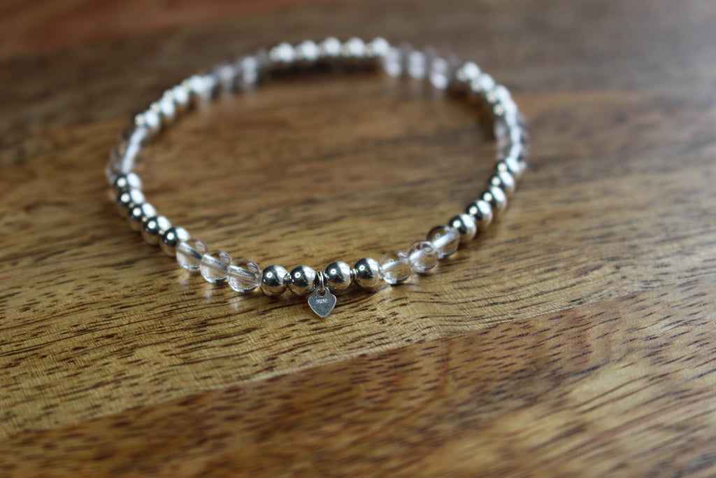 A beaded stretch bracelet with metallic silver beads, clear quartz beads, and a small silver heart charm, resting on a wooden surface.