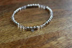 A beaded stretch bracelet with metallic silver beads, clear quartz beads, and a small silver heart charm, resting on a wooden surface.