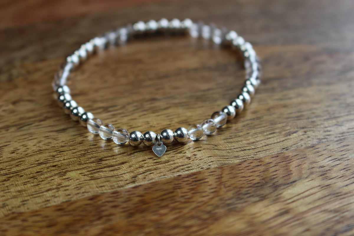 A beaded stretch bracelet with metallic silver beads, clear quartz beads, and a small silver heart charm, resting on a wooden surface.
