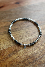 A beaded stretch bracelet with alternating black Hematite and silver beads, featuring a small silver heart charm, resting on a wooden surface.