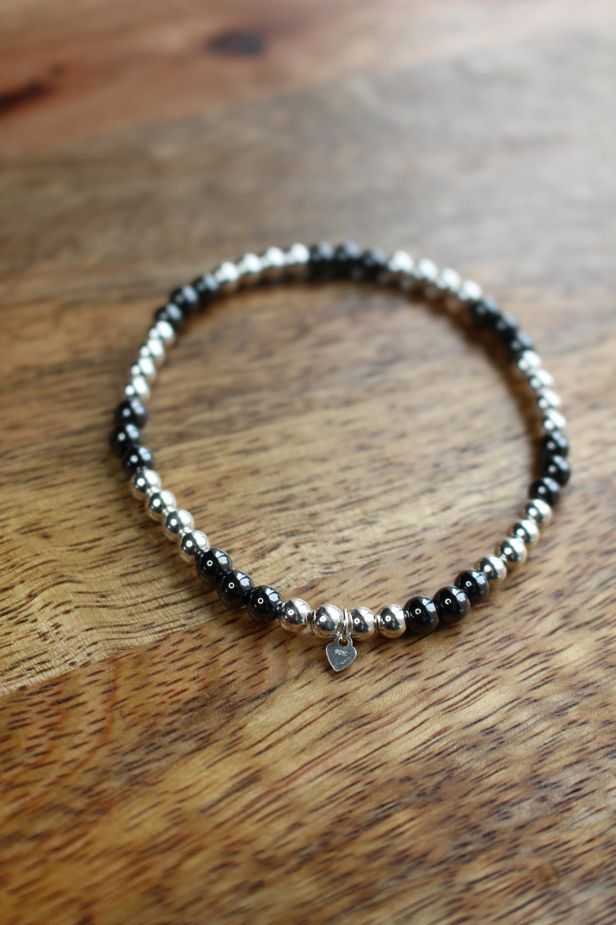 A beaded stretch bracelet with alternating black Hematite and silver beads, featuring a small silver heart charm, resting on a wooden surface.