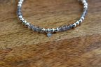 A beaded stretch bracelet featuring gray stone beads, polished silver metal beads, and a small silver heart charm, displayed on a wooden surface.