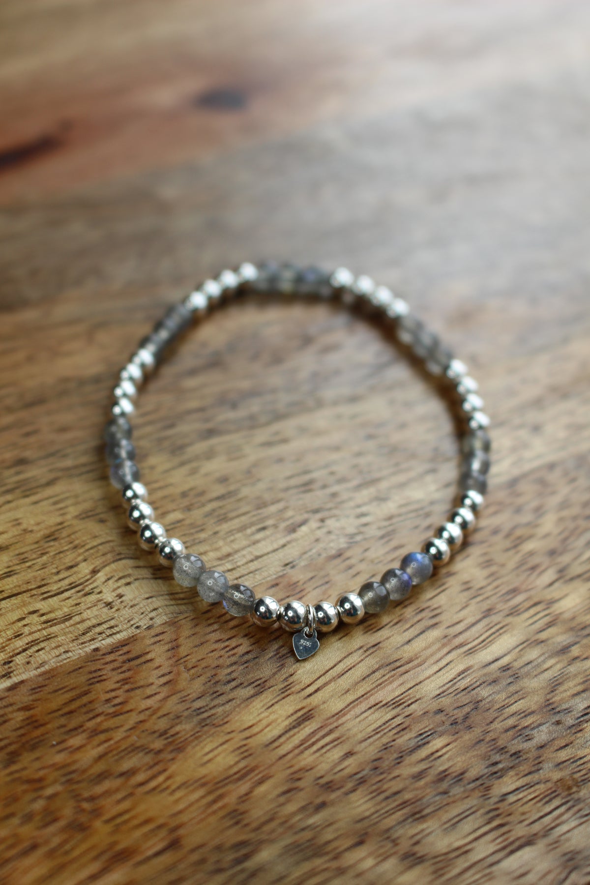 A beaded stretch bracelet featuring metallic silver beads, Labradorite beads, and a small silver heart charm, displayed on a wooden surface.