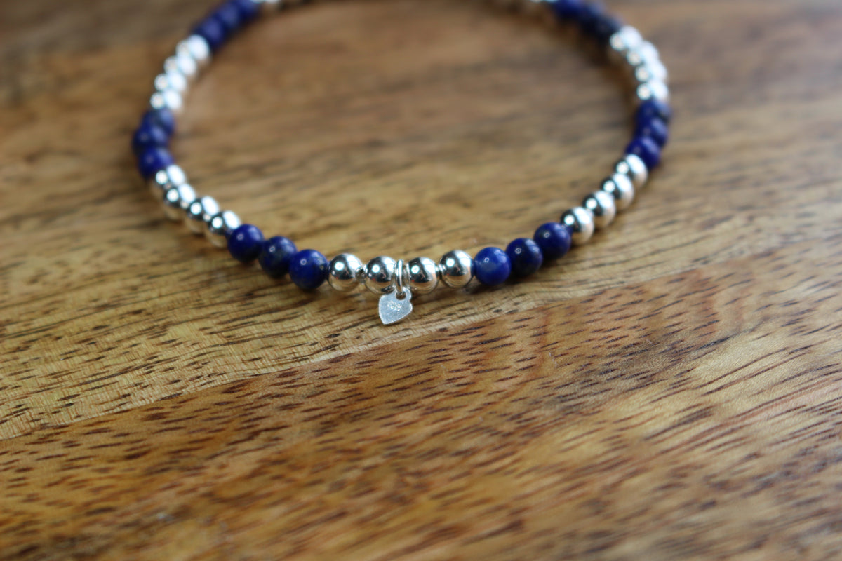 A beaded stretch bracelet featuring dark blue lapis lazuli and polished silver beads, with a small silver heart charm, resting on a wooden surface.
