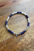 A beaded stretch bracelet with dark blue lapis lazuli and polished silver beads, featuring a small silver heart charm, displayed on a wooden surface.