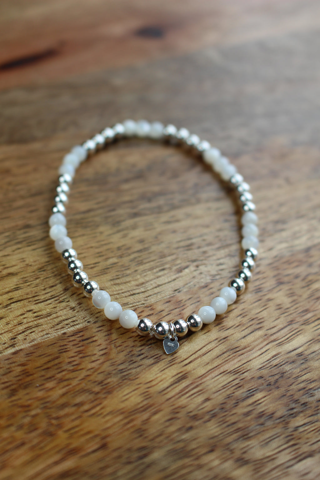 A beaded stretch bracelet featuring white round Mother of Pearl beads and polished silver spacer beads, with a small silver heart charm, displayed on a wooden surface.
