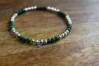 A beaded stretch bracelet featuring dark green malachite and silver beads with a small silver heart charm, resting on a wooden surface.