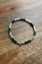 A beaded stretch bracelet featuring dark green malachite and polished silver beads with a small silver heart charm, displayed on a wooden surface.