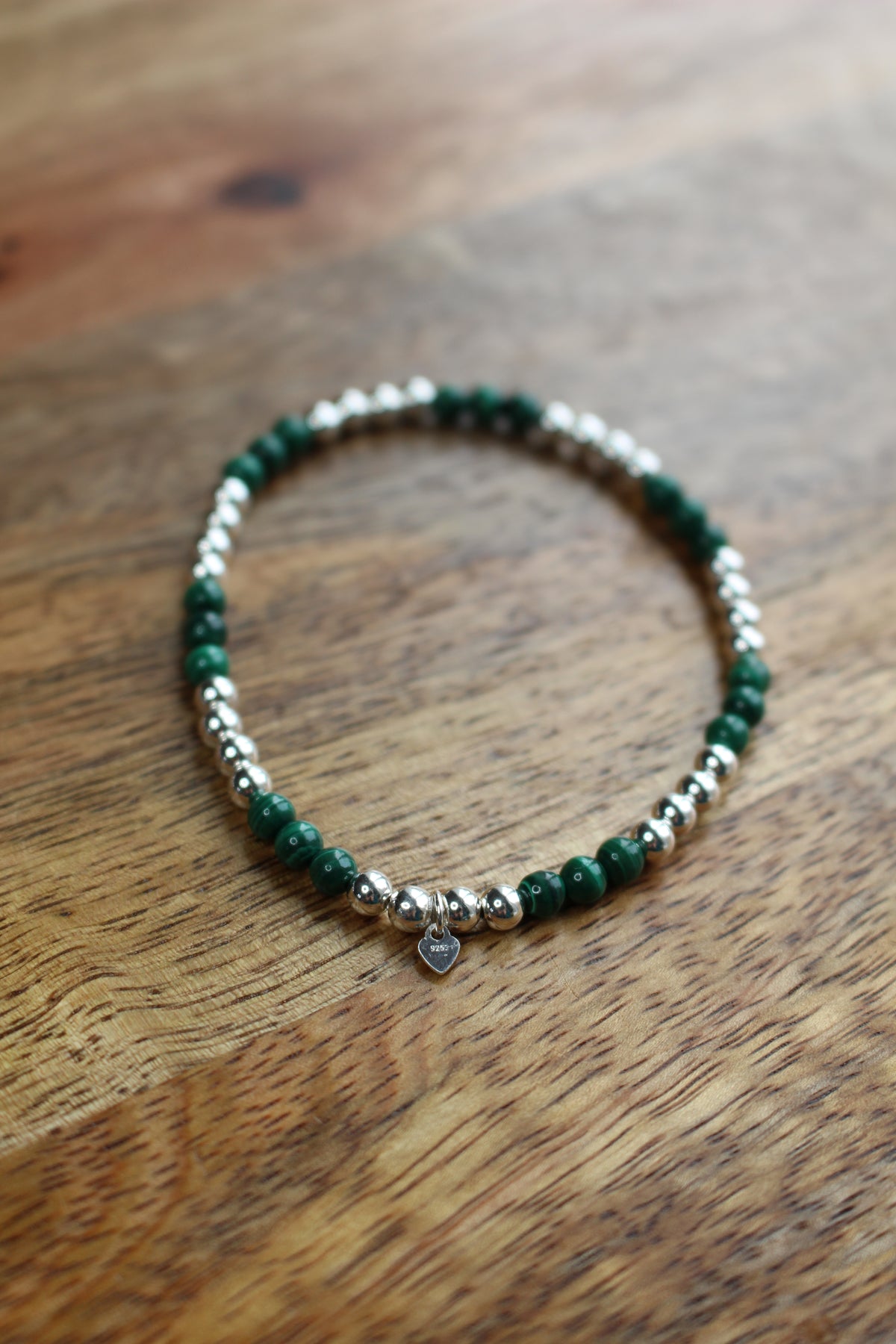 A beaded stretch bracelet featuring dark green malachite and polished silver beads with a small silver heart charm, displayed on a wooden surface.