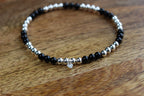 A beaded stretch bracelet with alternating black Obsidian and silver metallic beads, featuring a small clear heart-shaped charm, displayed on a wooden surface.