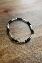 A beaded stretch bracelet with alternating black Onyx and silver beads, featuring a small silver heart charm, displayed on a wooden surface.