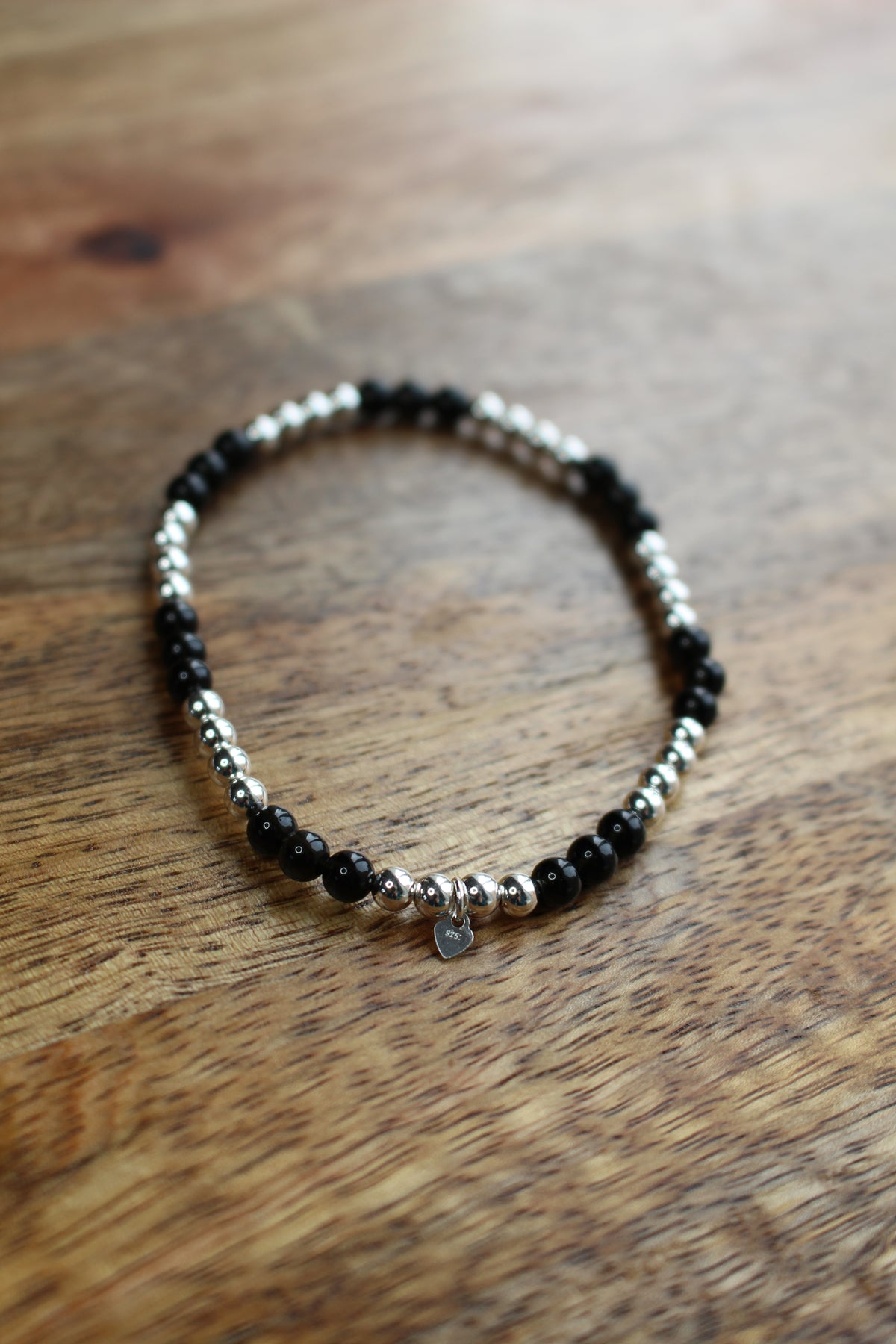 A beaded stretch bracelet with alternating black Onyx and silver beads, featuring a small silver heart charm, displayed on a wooden surface.