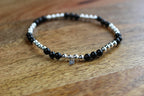 A beaded stretch bracelet with alternating black and silver metallic beads, featuring a small silver heart charm, displayed on a wooden surface.
