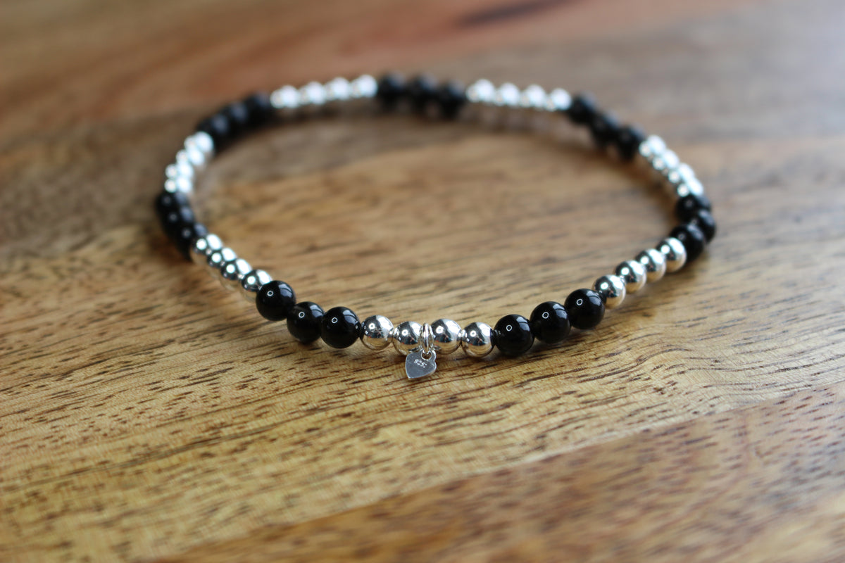 A beaded stretch bracelet with alternating black and silver metallic beads, featuring a small silver heart charm, displayed on a wooden surface.