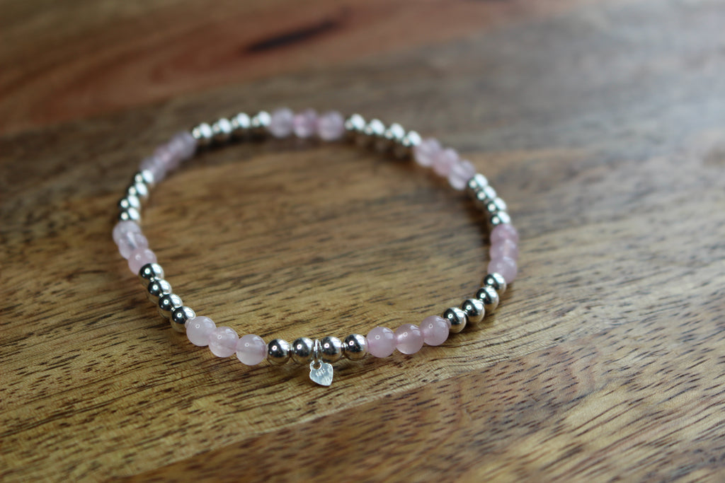 A beaded stretch bracelet featuring light pink rose quartz beads and polished silver spacer beads, with a small clear heart charm, resting on a wooden surface.