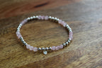 A beaded stretch bracelet featuring light pink rose quartz beads and polished silver spacer beads, with a small clear heart charm, resting on a wooden surface.