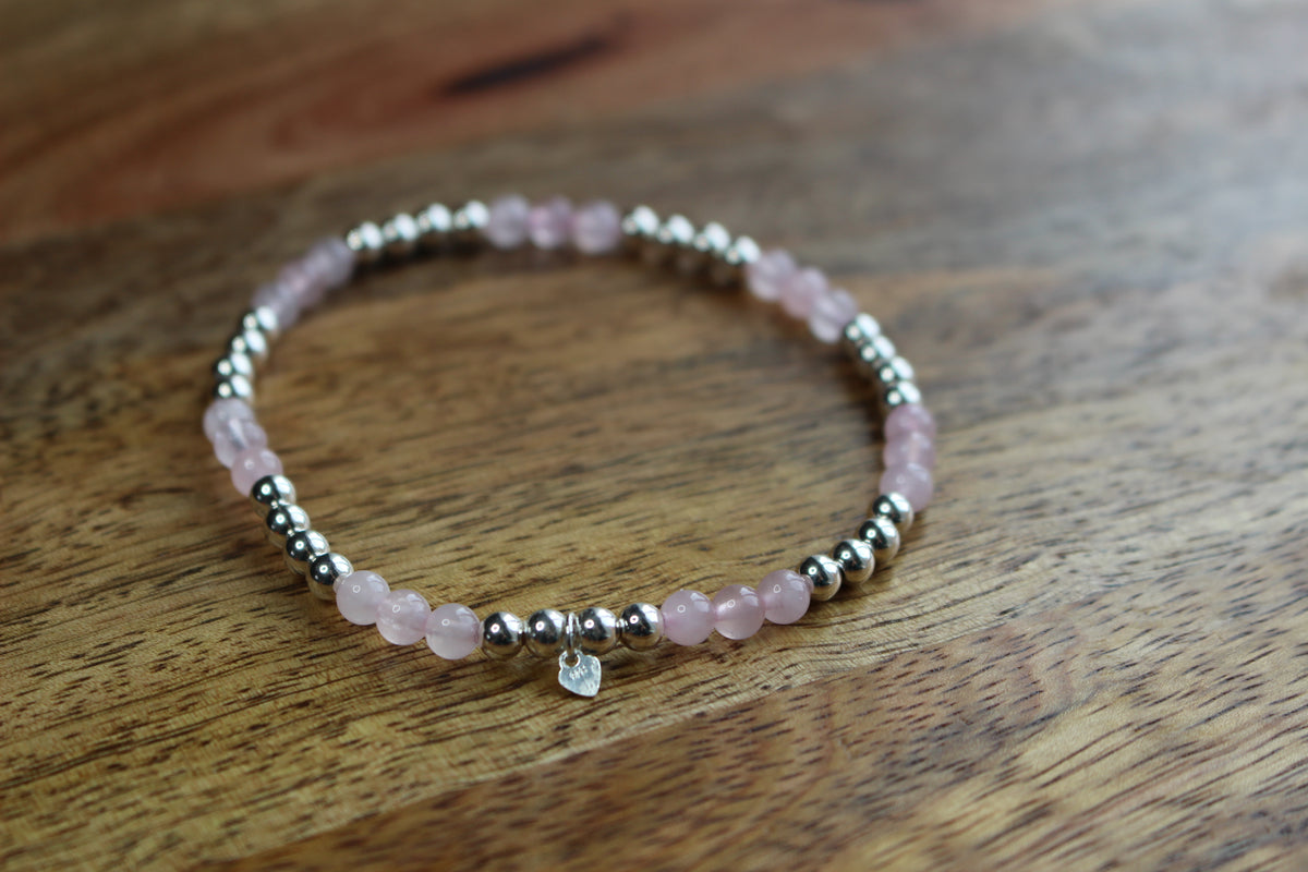 A beaded stretch bracelet featuring light pink rose quartz beads and polished silver spacer beads, with a small clear heart charm, resting on a wooden surface.