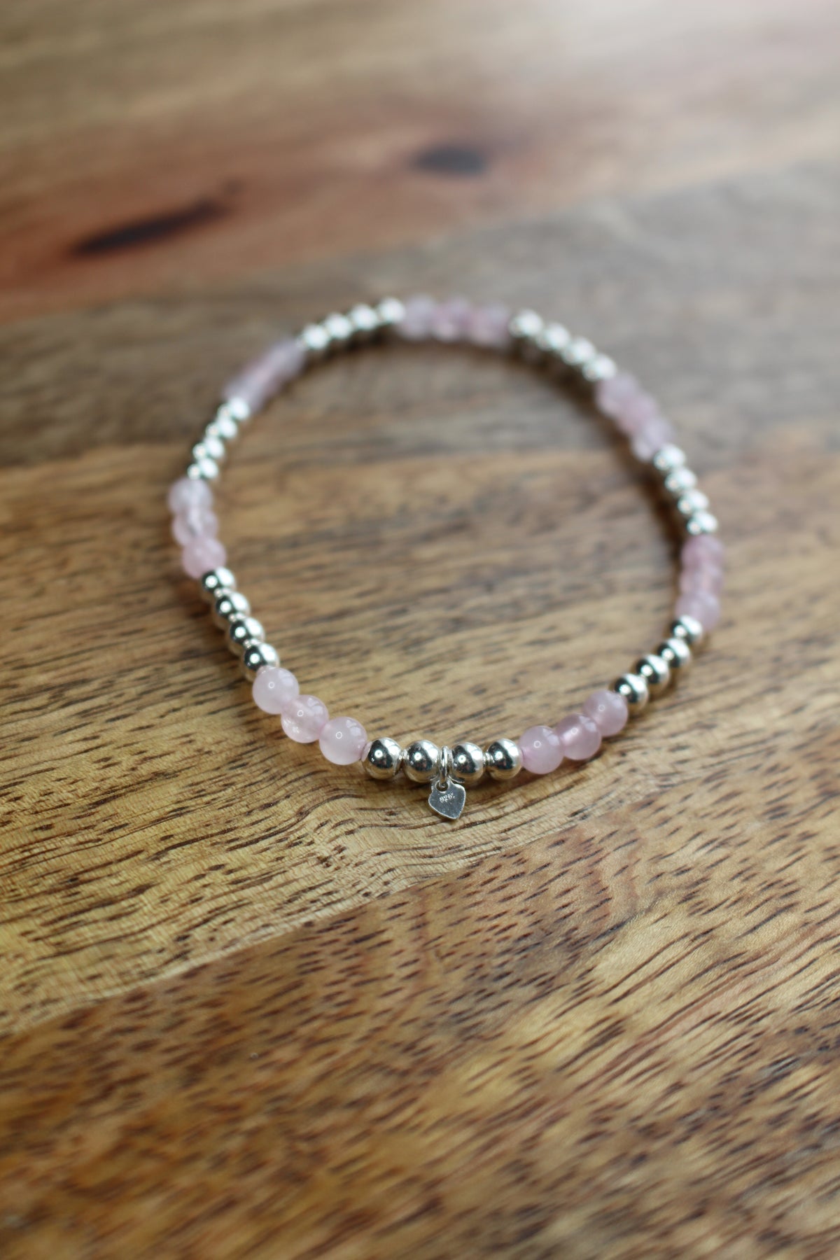 A beaded stretch bracelet with light pink Rose Quartz  beads, silver spacer beads, and a small silver charm, displayed on a wooden surface.