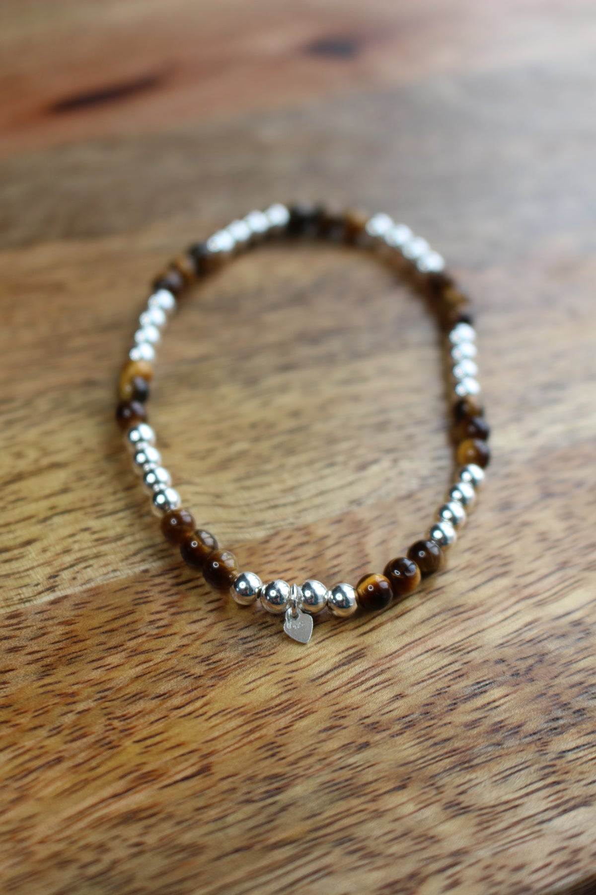 A beaded stretch bracelet with brown tiger's eye beads, silver spacer beads, and a small silver heart charm, resting on a wooden surface.