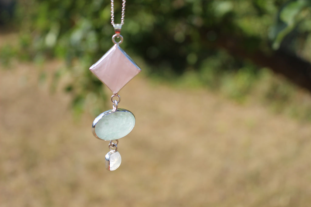 A silver pendant necklace featuring a pink diamond-shaped rose quartz stone, a light green Aventurine oval stone, and a small cushion moonstone, shown against a blurred outdoor background of green foliage and dry grass.